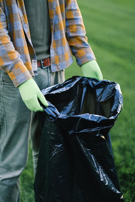A person standing outdoors on a grassy area, wearing a plaid shirt with yellow, blue, and gray tones, along with green gloves. They are holding open a large black plastic rubbish bag that is partially folded over at the top, ready for waste disposal. The person's midsection is visible, showing their jeans and a brown belt. The background features a blurred patch of vibrant green grass, indicating a natural environment suitable for waste collection or garden clearance activities. The scene suggests the person is engaged in an outdoor rubbish removal task, possibly related to garden waste or general refuse collection, with no visible waste inside the bag at this stage. The lighting appears natural, highlighting the textures of the gloves, plastic bag, and clothing material, supporting imagery pertinent to independent waste handling and rubbish disposal services.