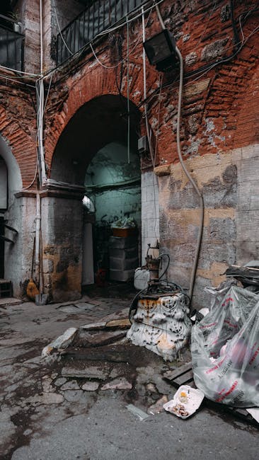 A narrow alleyway situated between a dark green corrugated metal building on the left and a tall, weathered brick wall on the right, with graffiti markings. The alley contains scattered debris and trash bags on the dirt ground, leading towards a brighter background with bare tree branches and a cloudy sky. Visible piping and electrical conduits run along the exterior wall of the building. An outdoor wall-mounted light fixture is attached above the alley, casting a faint glow. The scene suggests an urban environment where private waste clearance might be necessary, and the presence of waste and clutter highlights the importance of rubbish removal services, such as those offered by Waste Clearance Kentish Town, for efficient cleaning and disposal.