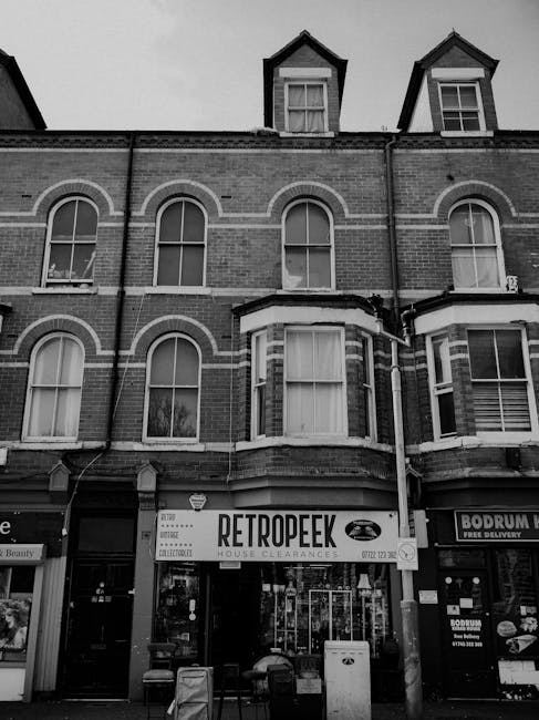The black and white photograph depicts a multi-storey brick building with a retail unit on the ground floor, identified by a large sign reading 'RETROPEEK' for house clearances. The upper floors feature several large, arched and rectangular sash windows with white frames and sills, some with partially visible curtains. The building's façade includes decorative brickwork detailing, and two small dormer windows with pitched roofs project from the roofline. In front of the retail space, there is an entrance with glass doors, and slightly to the right, another storefront displays a sign for 'BODRUM' offering free delivery. Various items, possibly collection bins or waste containers, are positioned on the pavement directly outside the retail units, suggesting additional waste or clearance activity in the vicinity. The overall scene captures an urban retail environment with an emphasis on private waste clearance services, indicative of alternative rubbish management options outside local authority collection. The photograph's neutral lighting and monochrome tone highlight the detailed textures of brickwork and window frames, contributing to a professional and observational portrayal of a building involved in waste clearance activities within a commercial setting.