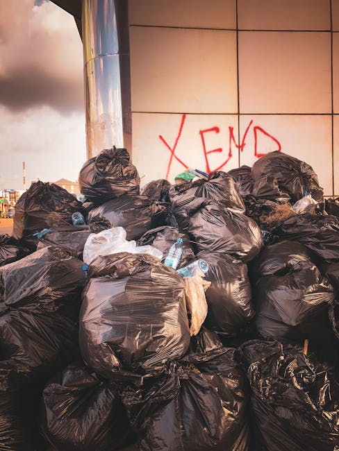 A pile of discarded cardboard boxes, some flattened and some partially collapsed, rest against a rough, weathered brick wall outdoors. The boxes vary in size, with some open and others sealed, made of plain brown and corrugated cardboard, displaying printed barcodes and labels. Among the boxes are crumpled paper and plastic packaging, with a white sack or bag partially visible beneath the refuse. The scene is set on a dirt surface with small stones and debris scattered around, bordered by a brick wall and a large tree trunk on the left side. Natural daylight illuminates the scene, casting soft shadows and highlighting the textures of the cardboard and ground. This image, associated with waste clearance services, illustrates a typical example of rubbish collection or on-site clearance handled by Waste Clearance Kentish Town, showcasing a typical accumulation of domestic or commercial waste awaiting removal.