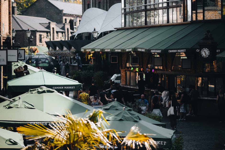 A busy outdoor scene at Camden Market featuring several large green umbrellas with the words 'Camden Market' printed on their edges, providing shade for numerous pedestrians walking along the street. In the background, there are brick buildings with large windows and a modern white canopy structure. The street is filled with people engaged in browsing or socialising, some standing and others walking. To the left, a black vehicle is partially visible behind the umbrellas, and there are planters with green foliage adding to the café or market atmosphere. The lighting suggests daytime with bright, natural sunlight illuminating the scene, creating a lively and vibrant environment typical of a busy market area. The overall setting emphasizes an urban commercial outdoor space, relevant in the context of waste management and the importance of managing rubbish generated in such popular public areas, where external private waste collection services may be used alongside or instead of council-provided rubbish disposal.