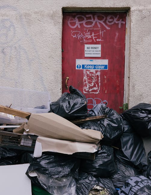 A pile of black rubbish bags and discarded cardboard boxes and packaging materials are overflowing in front of a red metal door with graffiti and multiple signs, including one reading 'No Money Back' and another with the 'Keep Clear' symbol. The bags and debris are placed on a pavement area adjacent to a beige, textured wall, with some items leaning against the door frame. The scene suggests local waste or fly-tipped rubbish in an urban environment, highlighting the need for independent rubbish removal services, such as those offered by Waste Clearance Kentish Town, to manage waste efficiently and maintain cleanliness in the area.