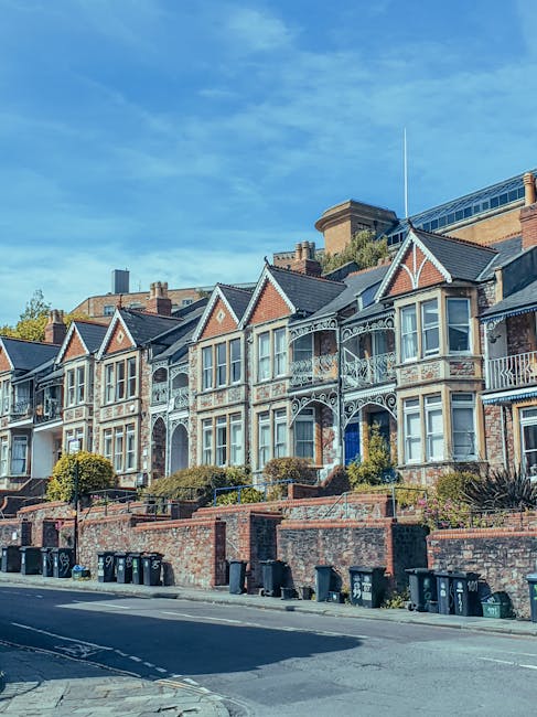A row of Victorian-style terraced houses with ornate white metal balconies and large bay windows, situated on a slightly elevated brick front garden level, with small trees and bushes in front. The houses are constructed of red brick with contrasting white window frames and decorative trim, featuring gabled roofs with dark slate tiles. In the foreground, a paved street runs parallel, lined with black wheelie bins positioned near the curb for waste collection. The sky above is clear with a bright blue tone, and a modern building with large glass panels is visible in the background behind the houses. This scene captures a typical residential street, where discreet waste management solutions like independent rubbish collection services may be necessary, especially for managing household waste in such period properties. Waste Clearance Kentish Town offers regular rubbish removal in the area, supporting private waste handling without reliance on local authority services, aligning with the context of on-site clearance and alternative waste disposal practices evident in the scene.