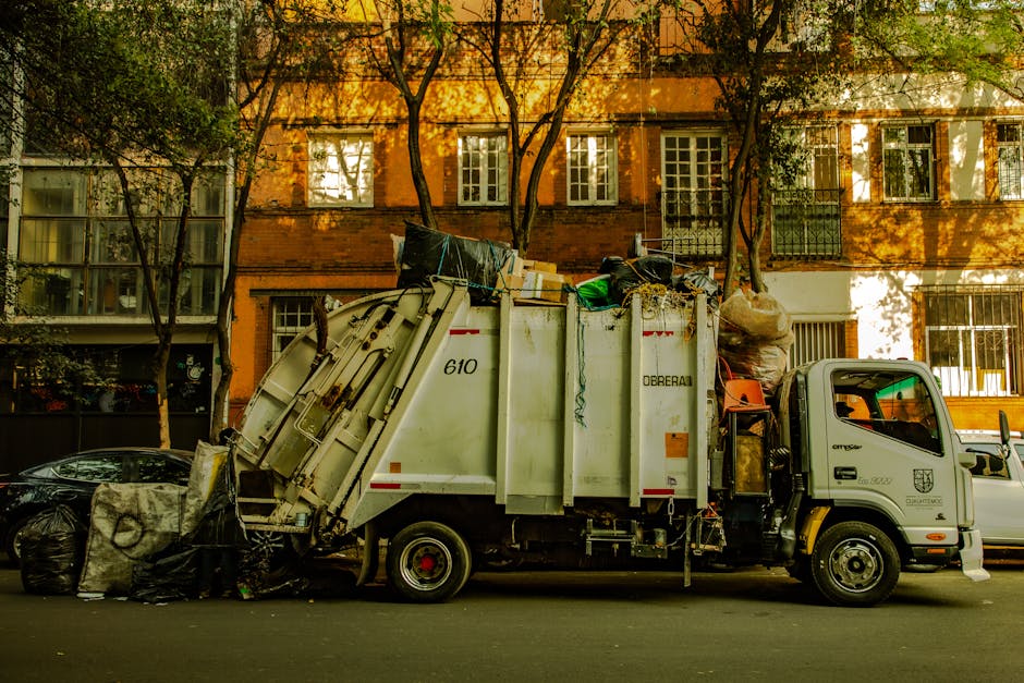 A white waste collection truck with the number 610 and company branding 'DEREAL' on its side is parked on a city street in front of a residential building with a warm orange brick facade and multiple windows, some with white frames and shutters. The truck's rear is open, revealing a load of mixed waste, including black garbage bags, cardboard boxes, and various discarded household items piled on top. Several large black trash bags sit on the pavement beside the truck, one of which appears slightly torn or open. The scene is shaded by tall trees with green leaves, casting dappled sunlight onto the building and street, creating a calm yet utilitarian atmosphere. This setup suggests an off-site or private waste collection service, such as those provided by Waste Clearance Kentish Town, for clearing rubbish from residential or commercial properties in the area.