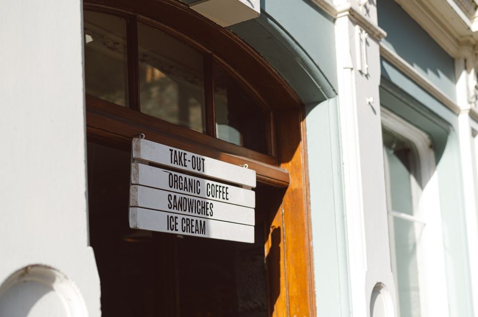 A close-up photograph of a white wooden signboard with black capital letters, hanging outside a café with a curved wooden frame window. The sign lists four items: 'Take-Out,' 'Organic Coffee,' 'Sandwiches,' and 'Ice Cream,' each on a separate line, with the sign angled slightly downward. The café's exterior wall is painted light pastel green, and nearby are decorative white molding details. The natural sunlight casts soft shadows on the wall and illuminates the sign, enhancing the textures of the wood and paint. The scene captures a quiet street atmosphere, with the sign serving as a subtle visual element related to service areas that might require waste or rubbish removal, typically handled by professionals such as Waste Clearance Kentish Town. The environment is clean and well-maintained, emphasizing an orderly setting suitable for waste clearance or disposal activities.