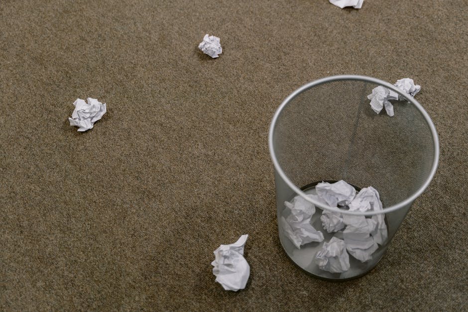A cylindrical transparent waste bin made of clear plastic is positioned on a beige carpeted floor, filled with scattered crumpled pieces of white paper. Several additional crumpled papers are strewn across the floor around the bin, some lying close to it and others further away, illustrating a scene of paper waste or discarded notes. The paper objects have varying sizes, with visible folds and creases, and their white color contrasts against the textured, light brown carpet. The environment appears to be an indoor office or workspace setting, with an emphasis on waste disposal in a private or independent context, as managed by waste clearance services such as Waste Clearance Kentish Town. The lighting is neutral, evenly illuminating the scene and highlighting the scattered rubbish, indicating a typical clean-up or rubbish collection scenario that aligns with the context of rubbish removal and disposal.