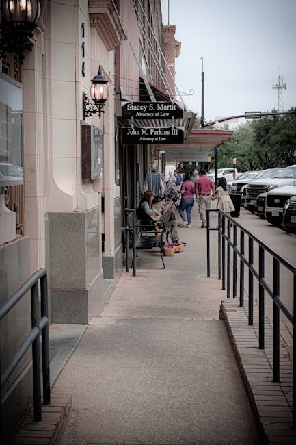 A view of a busy city sidewalk in front of a row of commercial buildings with a storefront sign indicating legal services. In the foreground, a sidewalk features a black metal railing along the right side, leading towards the background where several pedestrians are walking and sitting. Some people are standing near a bench with a person seated, possibly waiting or resting, and a woman in a pink jacket is seen walking past. Parked cars line the street on the right, with visible vehicle tops and windshields suggesting parking spaces for private vehicles. The buildings have decorative elements such as lantern-style lights attached to the exterior wall and signage for legal offices, including Stacey S. Martin and John M. Perkins III, attorneys at law. The environment appears overcast, with soft, diffuse lighting typical of a cloudy day. This scene may be typical of a commercial district where businesses and professional services are accessible to pedestrians, aligning loosely with local rubbish removal or waste collection needs in urban areas managed by companies like Waste Clearance Kentish Town, ensuring hygienic and clutter-free environments for businesses and visitors alike.