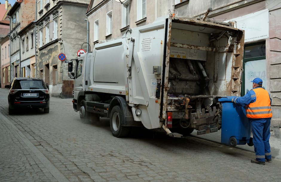 A white refuse collection truck operated by Waste Clearance Kentish Town is parked on a narrow urban street with cobblestone pavement, positioned beside a sidewalk adjacent to multi-story residential buildings with weathered facades. The truck's rear loading compartment, which shows signs of rust and wear, is open and appears to be in the process of collecting waste or rubbish from the street or nearby areas. A worker dressed in a high-visibility orange vest and blue uniform is standing to the right of the truck, placing or retrieving a blue recycling bin into or from the truck's rear compartment. The street scene is highlighted by parked cars, including a black vehicle with a UK license plate, and various street signage, including no-parking or stopping signs. The environment reflects an everyday urban setting where private waste collection services, such as those provided by Waste Clearance Kentish Town, facilitate rubbish removal outside of local authority collection routes, especially in residential or commercial areas with limited space for waste storage.