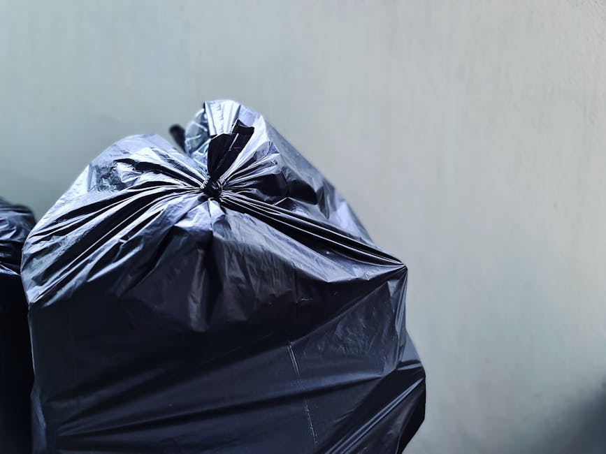 Close-up of a tightly tied black rubbish bag made from glossy plastic material, positioned in the foreground against a plain, light-colored wall. The bag is resting on an outdoor surface such as a driveway or pavement, with its top gathered and sealed, indicating it is ready for collection. Additional similar black rubbish bags are partially visible behind it, suggesting a collection of waste awaiting removal. The minimal background and natural lighting highlight the texture and sheen of the plastic material, within an environment consistent with private waste disposal. The scene aligns with private rubbish collection services, such as those offered by Waste Clearance Kentish Town, for on-site waste clearance and alternative disposal methods outside local authority services.