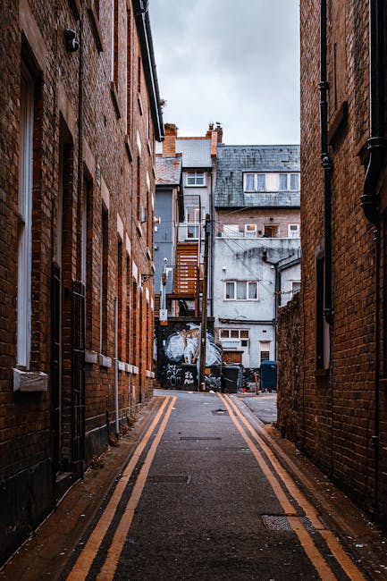 A narrow urban alleyway between two red brick buildings, with a paved surface featuring double yellow lines along each side. The alley leads towards a small courtyard at the back, where several large black trash bins are visible, placed against a plain, weathered grey wall. Above the bins, a wooden staircase with a metal railing ascends to an upper floor of an adjoining building. The surrounding brick walls are textured, with visible mortar lines and some minor weathering. The lighting is diffuse, suggesting an overcast sky, and the environment appears clean but cluttered with waste collection containers, indicative of private rubbish handling services such as those offered by Waste Clearance Kentish Town. The scene captures an outdoor space where alternative waste removal methods are evident, contributing to the context of efficient rubbish removal in a densely built urban setting.