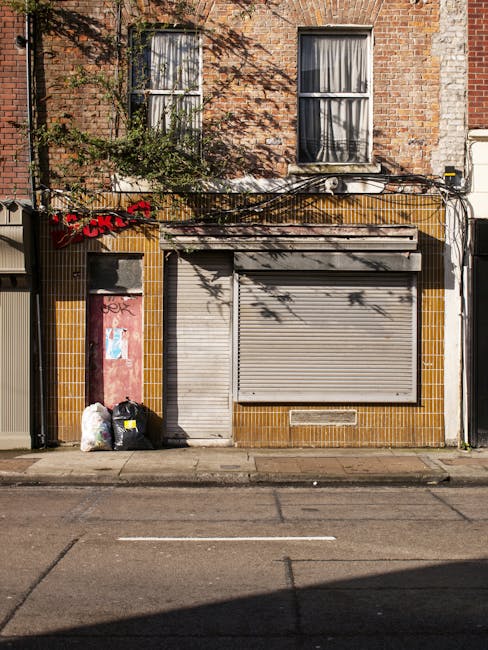 A brick building with two vertically aligned windows featuring white frames and partially drawn curtains on the upper floor. The ground floor has a metallic roller shutter door, painted in a light, weathered grey, covering a shop or commercial unit. To the left of the shutter, there is a narrow doorway with a red slab or panel beside it, which appears weathered and slightly graffitied. Two black plastic rubbish bags and a transparent bag filled with waste are placed against the building's wall near the doorway, indicating private waste disposal. The building's facade is finished with yellowish-brown tiles around the lower section, contributing to a worn urban appearance. Above, there is a small air conditioning unit, with shadows from nearby trees cast across the facade, adding contrast. Electrical wires and cables run horizontally across the building's upper section, some attached to the wall, reflecting common urban infrastructure. The street in front of the building is paved with asphalt, showing faint lane markings, suggesting an accessible location for rubbish clearance and alternative waste handling services offered by Waste Clearance Kentish Town.