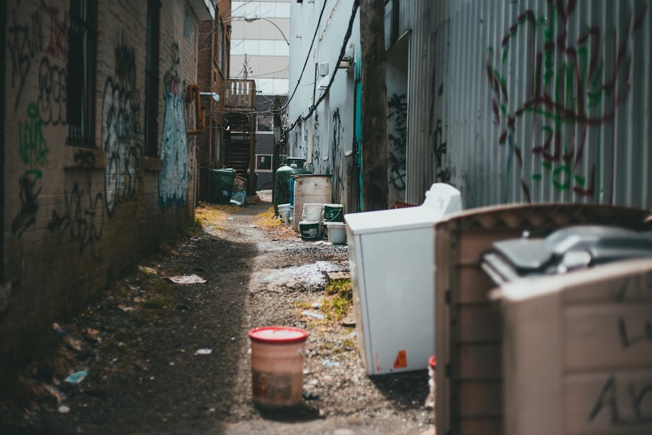 A narrow urban alleyway between two red brick buildings, with a paved surface featuring double yellow lines along each side. The alley leads towards a small courtyard at the back, where several large black trash bins are visible, placed against a plain, weathered grey wall. Above the bins, a wooden staircase with a metal railing ascends to an upper floor of an adjoining building. The surrounding brick walls are textured, with visible mortar lines and some minor weathering. The lighting is diffuse, suggesting an overcast sky, and the environment appears clean but cluttered with waste collection containers, indicative of private rubbish handling services such as those offered by Waste Clearance Kentish Town. The scene captures an outdoor space where alternative waste removal methods are evident, contributing to the context of efficient rubbish removal in a densely built urban setting.