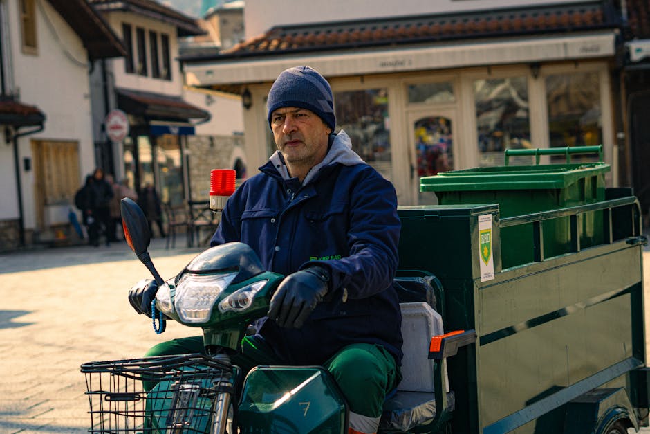 A man wearing a dark blue jacket, gray hoodie, and navy beanie is seated on a three-wheeled electric cargo bike with a large green waste container attached to the rear. The container has a hinged lid and is secured to the bike’s frame, designed for waste collection or rubbish removal services. The man is holding the handlebar with black gloves, situated on a paved outdoor area in a town or shopping district. In the background, there are storefronts with glass windows, some with signage, and a few pedestrians walking along the street. The scene is illuminated by natural daylight, casting soft shadows on the ground. This setting illustrates a typical city or town environment where independent waste collection or rubbish removal, facilitated by service providers like Waste Clearance Kentish Town, takes place, emphasizing eco-friendly and efficient waste management in local communities.
