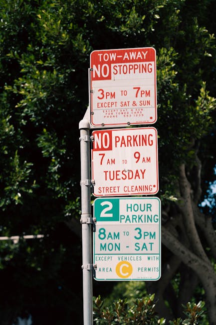 A multi-layered street sign post positioned outdoors against a backdrop of green leafy trees and a blue sky. The top sign is red with white text, indicating a tow-away zone with no stopping rules from 3pm to 7pm, excluding Saturdays and Sundays, and providing a contact phone number. Below it, a second red sign states no parking is permitted from 7am to 9am on Tuesdays for street cleaning. The third sign is a green and white parking restriction indicating a two-hour parking limit from 8am to 3pm, Monday to Saturday, with a note that it applies to vehicles with a valid area C permit. The signs are mounted on a metallic pole, which is partly visible, and appear to be in a maintained urban area. The lighting suggests daylight conditions, highlighting the clarity of the signage while the leafy background provides a natural contrast. The scene subtly relates to regulations surrounding private or independent waste management activities, which may involve compliance with local parking and stopping rules when managing rubbish clearance or disposal tasks in the vicinity.