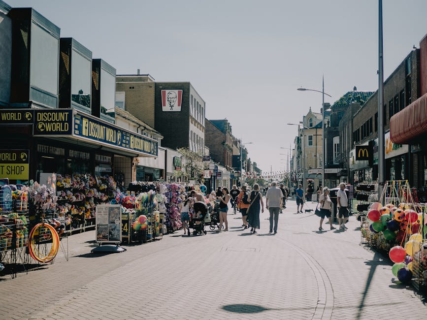 A bustling street scene in a commercial area with shops on both sides, including a store with a bright yellow sign reading 'Discount World' and other storefronts such as a McDonald's and a fashion retailer. The street is populated with pedestrians, some walking, others pushing strollers, and a few carrying shopping bags. On the left side, outdoor stalls display an array of colorful goods, including flowers, balloons, and toys, arranged on tables and racks. The pavement is light-colored and textured, with faint tram or trolley markings visible on the street surface. The buildings are multi-story, with varying architectural styles, some featuring large window displays and signage. Bright natural lighting suggests a clear day, with minimal shadows cast by the pedestrians and street fixtures. The scene captures a lively urban environment consistent with busy shopping districts, where independent collection or on-site clearance of waste or unwanted items may be necessary for vendors and shoppers, naturally aligned with private waste handling services such as those provided by Waste Clearance Kentish Town.